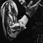 A man in a waistcoat smoking a cigarette, shirt sleeve covered in blood.